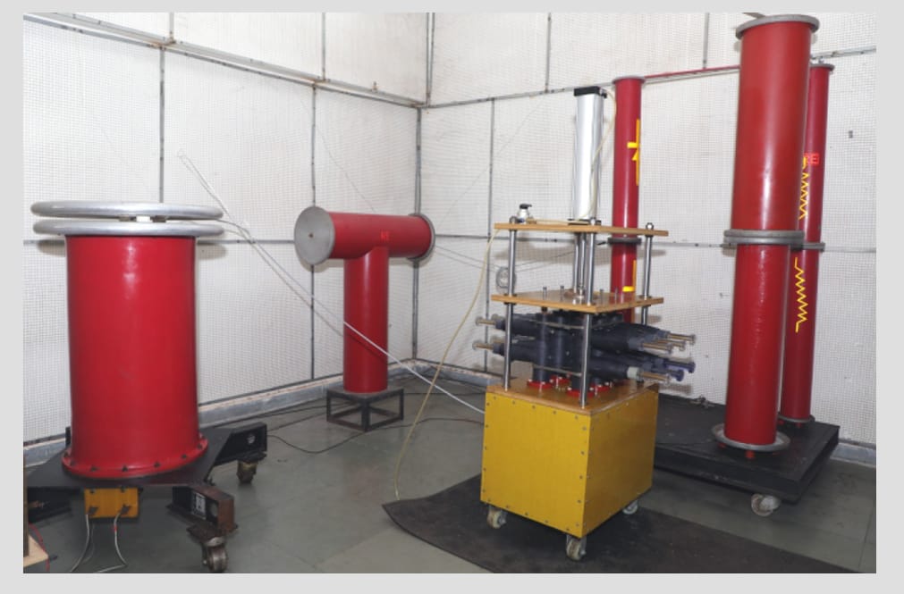 Laboratorio de alta tensión del fabricante: dos transformadores de ensayo cilíndricos rojos y un divisor capacitivo, con tres terminaciones montadas sobre una base amarilla, dentro de un recinto apantallado con malla.